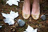 Close up of a woman's feet wearing golden shoes standing on a forest floor with silver leaves