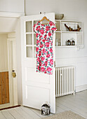 Pink floral dress hanging on an open bedroom door beside vintage radiator and wall unit filled with bottles and jars and figurines