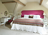 Striped bed cover on bed with pink headboard in beamed attic bedroom conversion Gloucestershire farmhouse, England, UK