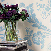 Detail of flower display in a glass vase resting on a stack of CD's with blue and white floral patterned wallpaper on walls