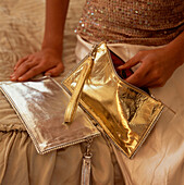 Woman dressed in eveningware sitting on a bed opening a gold fabric handbag