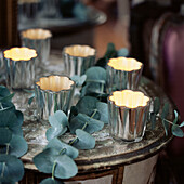 Gruppe von sechs silbernen Glas-Teelichthaltern mit leuchtenden Kerzen auf einem rustikalen Beistelltisch