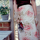 Stehende Frau mit Gartengeräten in einem Gartenhaus und einer geblümten Schürze in der Hand