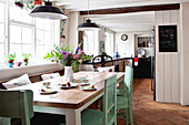 Black pendant lamps hang above kitchen table with green painted chairs, set with tea and cake
