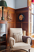 Cream armchair with glass-fronted cabinet in wood panelled Arts and Crafts style living room, Surrey, UK