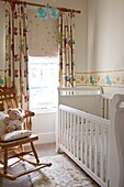 Weiß lackiertes Kinderbett mit Schaukelstuhl am Fenster in einem Einfamilienhaus, Großbritannien