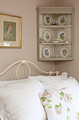 Decorative plates on wall mounted corner shelf above bed with floral cushion in semi-detached home UK