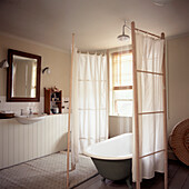 Duschkopf mit Wasser, das in die Badewanne fließt, ein hölzerner Paravent mit Duschvorhang in einem Badezimmer im Landhausstil