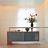 Anrichte mit Glasfront und Vase mit roten Rosen auf dem Treppenabsatz vor einem Fenster mit mattierte Plexiglasscheibe und einer seitlich beleuchteten Deckenplatte