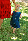 Rückansicht eines kleinen Mädchens mit besticktem Jeanskleid neben seiner Mutter im Garten