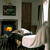 Cottage-Innenraum mit üppigen Decken auf der Chaiselongue vor einem beleuchteten gusseisernen Holzbrenner