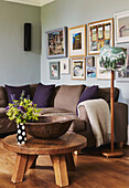 Artwork display above brown sofa with purple cushions and low wooden table in living room of family home, Rye, East Sussex, England, UK
