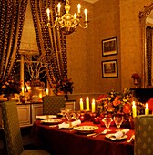 Beleuchtete Kerzen auf einem weihnachtlichen Esstisch mit goldenem Kronleuchter und zurückgebundenen gemusterten Vorhängen
