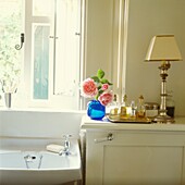 Bathroom sink below window with cut flowers and tray of perfumes with lamp 