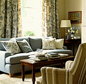 Light blue sofa with cushion co-ordinating curtains and striped armchair on opposite side of coffee table with books