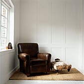 Brown leather armchair at uncurtained window with low ottoman stool in white panelled room