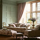 Floral patterned sofa an ottoman footstool in panelled room with large windows and checked curtains