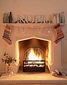 Single word 'NOEL with antique bottles and Christmas stockings on mantlepiece above lit fire