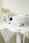 Bed with white bedlinen and embroidered floral cushions with bedside table