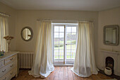 Full length cream curtains at French windows with coastal sea view from 16th century Rectory house Cornwall UK