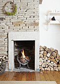 Stacked firewood and lit fire with exposed stone chimney breast in West Yorkshire living room  UK