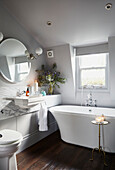Circular mirror above washbasin with freestanding bath below window in East London townhouse  England  UK
