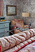 Vintage-Spiegel und Steppdecken mit Sessel in einem mit Metallblumentapeten tapezierten Schlafzimmer in einem Londoner Haus, UK
