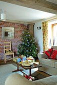 In Geschenke eingewickelte Geschenke auf dem Couchtisch im Wohnzimmer eines Hauses im ländlichen Suffolk mit einem Weihnachtsbaum vor einer freiliegenden Backsteinmauer, England, UK