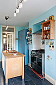 Wooden island unit with wall mounted shelving in blue kitchen of Bovey Tracey family home, Devon, England, UK