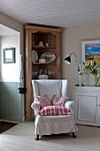 Striped cushions on slip-covered wingback armchair with wooden cabinet in corner of beach house Cornwall England UK