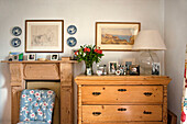 Family photographs on wooden chest of drawers with floral patterned chair at fireplace in cottage bedroom Cornwall UK