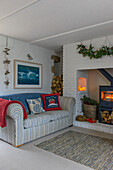 Gestreiftes Sofa mit blauer Decke unter einem gerahmten Kunstwerk mit beleuchtetem Holzbrenner zu Weihnachten in einem Bauernhaus in Penzance, Cornwall, England