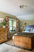 Wooden blanket box at foot of double bed with floral curtains at window in Helston farmhouse Cornwall UK