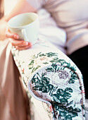 Entspannende Frau mit einer Tasse in der Hand auf der Armlehne eines geblümten Sofas