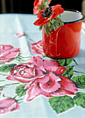 Rote Anenome in Emaille-Tasse auf Tischtuch mit Blumenmuster