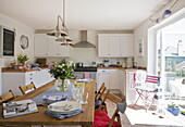 Wooden table in white fitted kitchen with open doorway to terrace, Dartmouth, Devon, UK