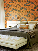 White leather stool at foot of double bed with orange floral wallpaper in London home, UK