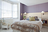 White painted chair at bay window with Venetian blinds in purple contemporary bedroom, fur throw over bed, London, UK
