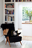 Tabby-Katze sitzt auf schwarzem Sessel mit Bücherregalen und Blick in die Küche in einem modernen Londoner Haus, UK