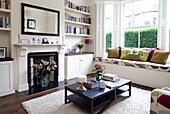 Recessed bookshelves with Victorian fireplace in living room with upholstered window seat in London townhouse, England, UK