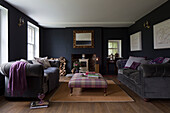 Tartan ottoman with grey velvet sofas in living room with carved wooden mirror and firewood in Sussex home UK