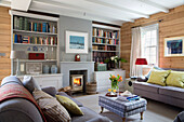 Light grey sofas in wood panelled living room with lit fire in Surrey home, England, UK