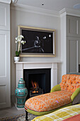 Orchid on mantlepiece with artwork and upholstered chaise longue at fireside in London bedroom, England, UK