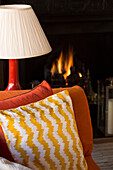 Patterned cushion and lamp in front of lit fire in London townhouse UK