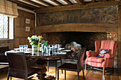 Upholstered armchair at inglenook fireplace with wooden dining table in timber framed Kent farmhouse UK