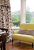 Yellow two-seater sofa in bay window with foliate patterned curtains Gloucestershire home England UK
