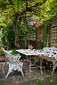 White wrought iron seating with table on patio below tree in back garden of London home UK