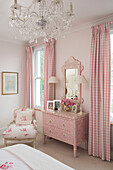 Pink gingham curtains and decorative chest of drawers with floral chair and chandelier in bedroom of Victorian terrace house South London UK