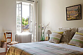 Striped pillows on checked bed at open window with chair in 19th century Provencal farmhouse France