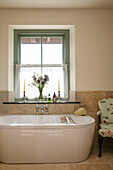 Geblümter Stuhl mit Badewanne unter einem Fenster mit Gardinen in einem Haus in Devon, Großbritannien